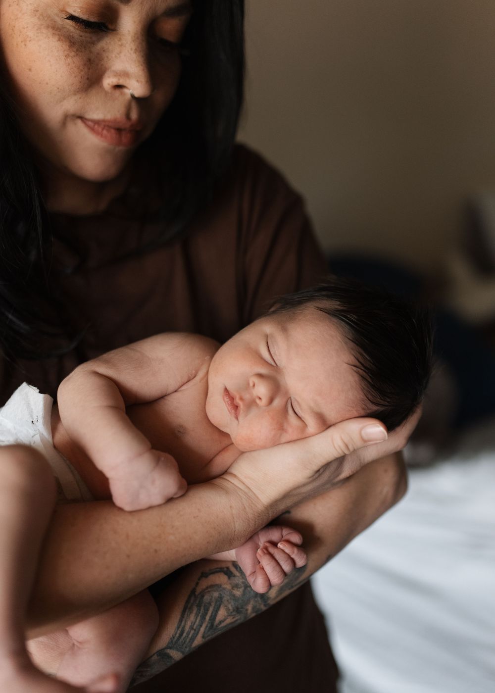 A tattooed mother tenderly holds her sleeping newborn baby against her chest in a warm, intimate portrait.