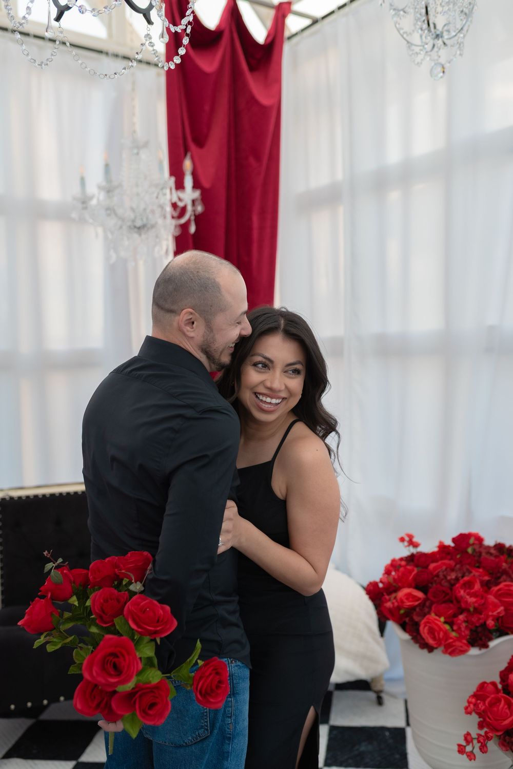 Romantic couple embraces near red roses and white curtains.