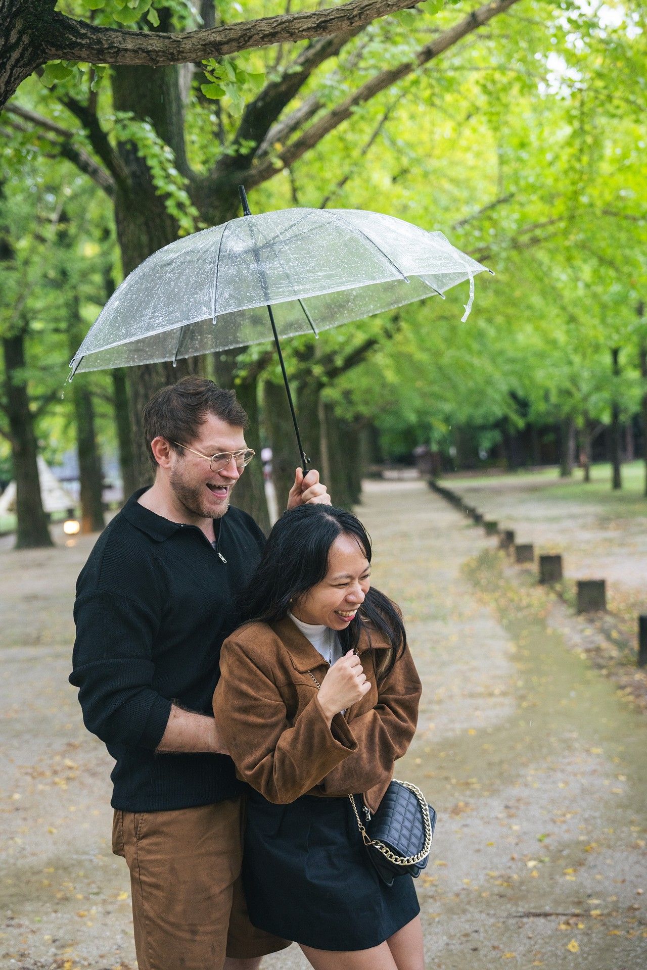 A couple shares a clear umbrella while laughing together on a tree-lined path during a rainy autumn day.
