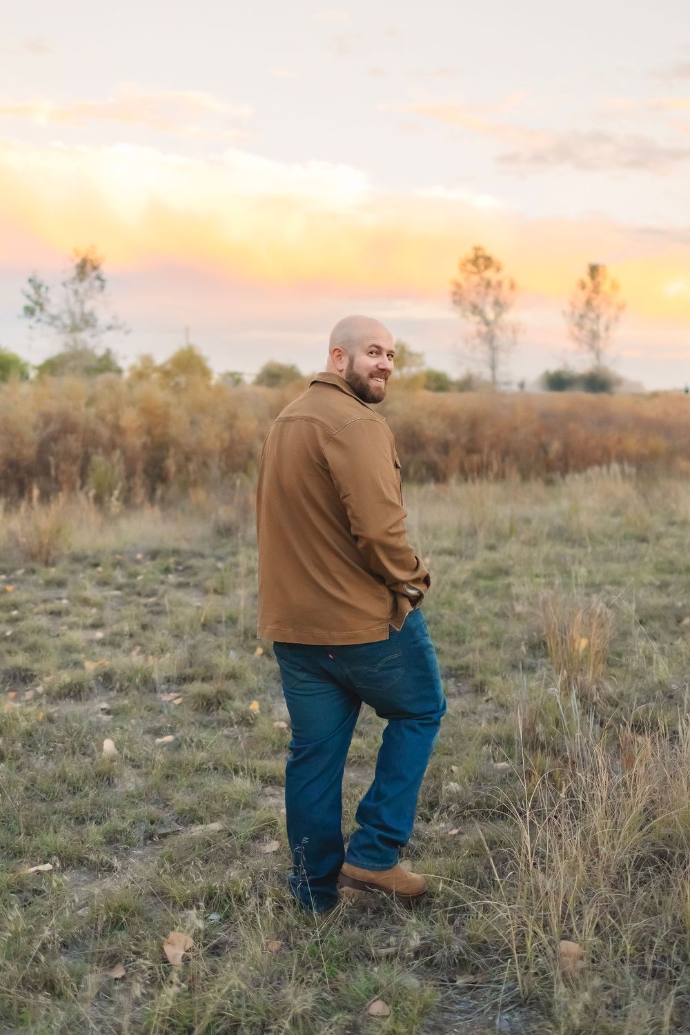 A man wearing a brown jacket and jeans stands in a grassy field during golden hour sunset.