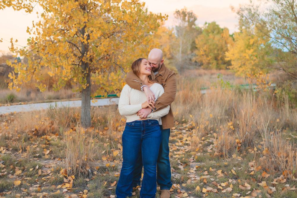 A couple embraces outdoors during autumn with golden leaves and natural scenery in the background.