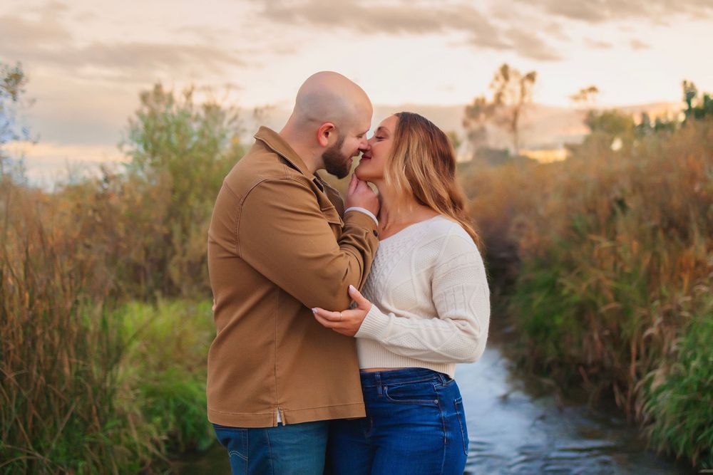 A couple embraces during a romantic sunset photoshoot near a winding river surrounded by tall grass and golden light.