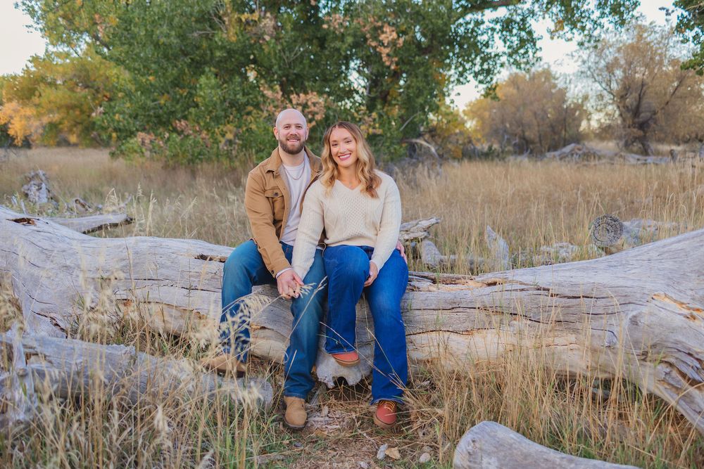 A couple sits together on rocks near dry grass and trees during a fall outdoor photo session.