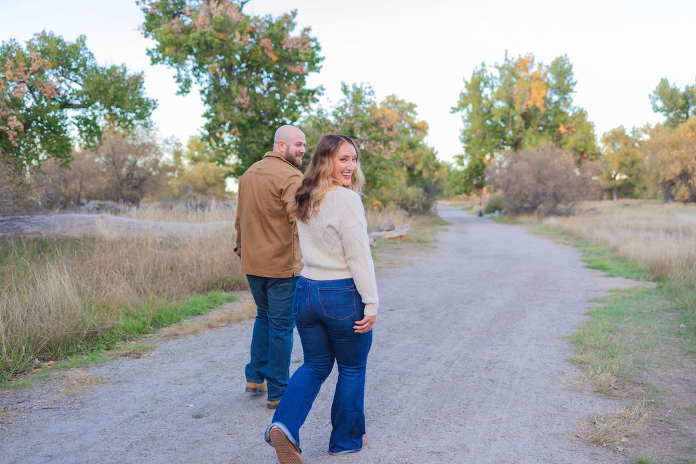 Couple walking together on a scenic dirt path surrounded by autumn foliage and tall grass at sunset.