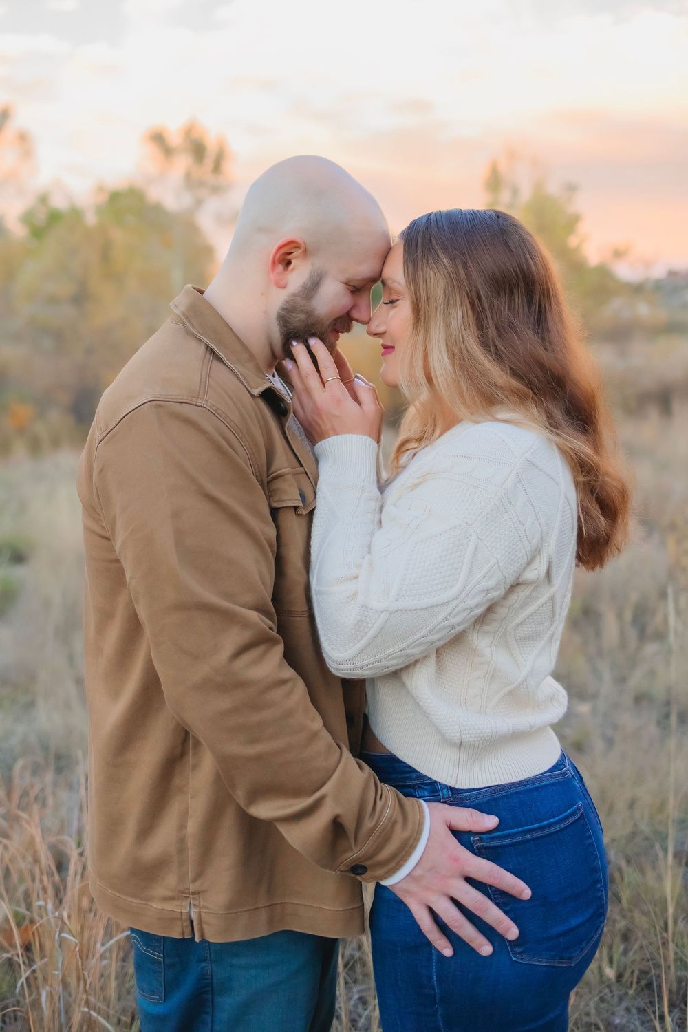 A couple embraces and kisses in a field during golden hour sunset, wearing blue jeans and tan outerwear.