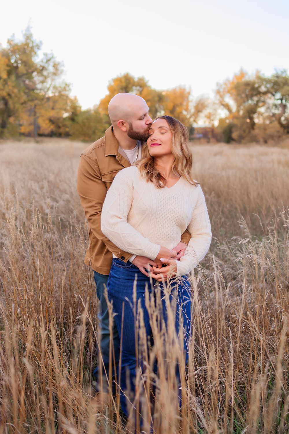 Romantic couple embraces in golden autumn field with tall grass during sunset photoshoot.