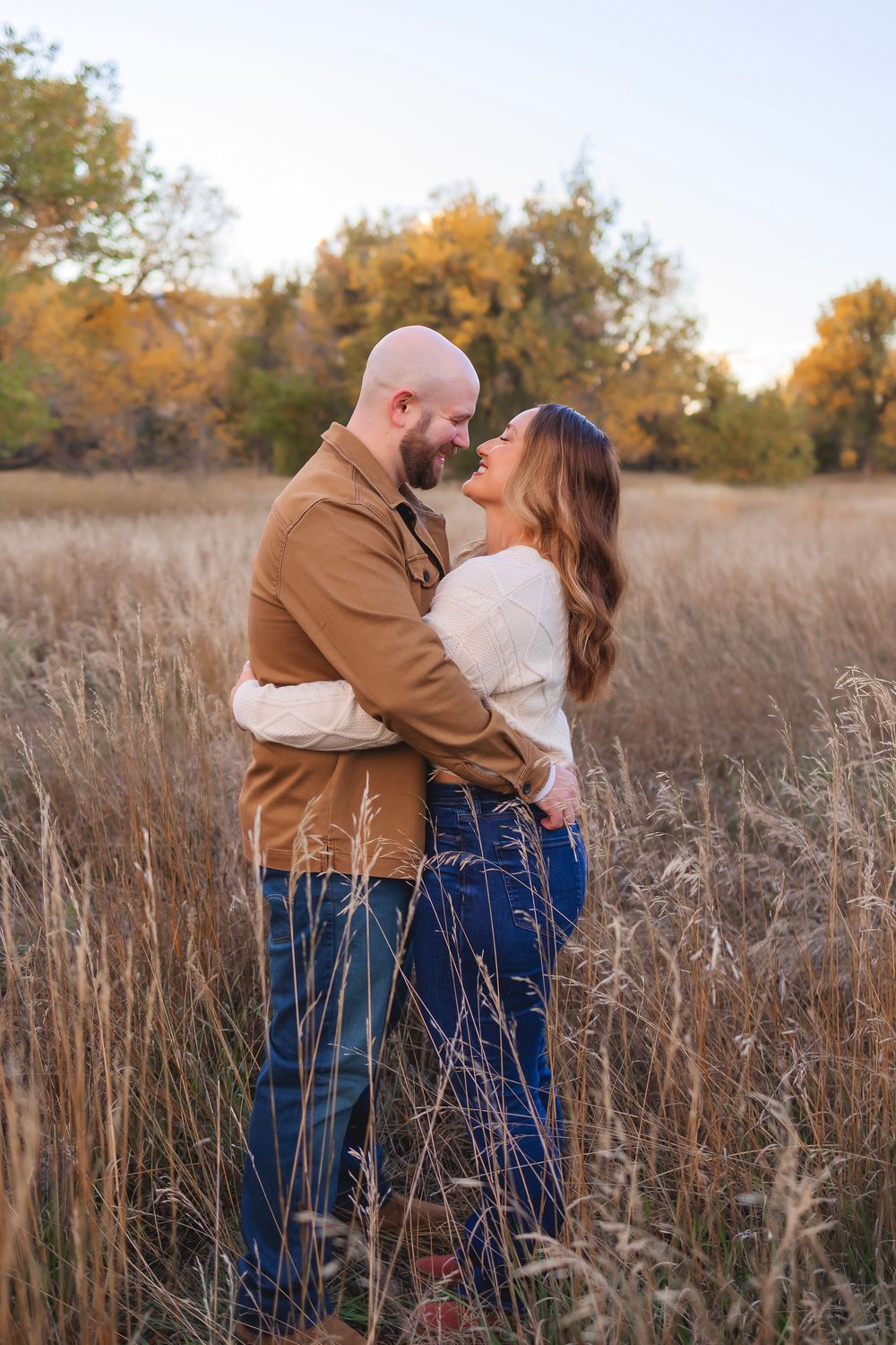 A romantic couple embraces in a golden autumn field with tall grass and fall foliage in the background.