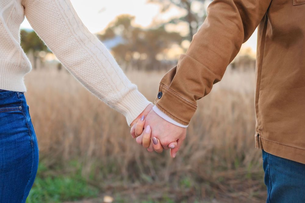 Two people hold hands while standing in a field with tall grass during golden hour.