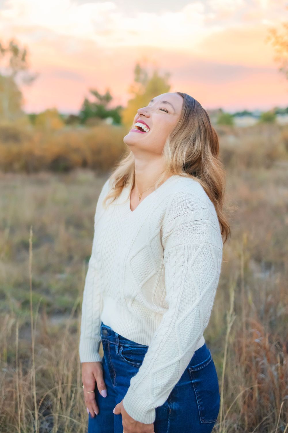 Person in white sweater and jeans laughs joyfully in golden field at sunset.