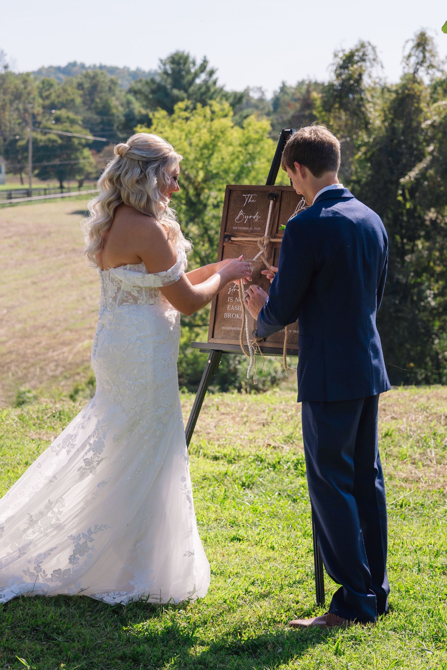 A couple in wedding attire doing a braiding ceremony together in a sunny outdoor field setting.