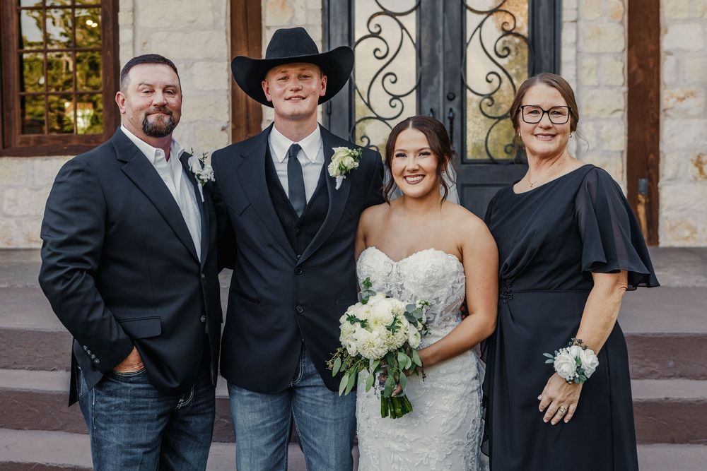 Wedding party members in formal attire pose together outside a stone building with ornate iron gates.