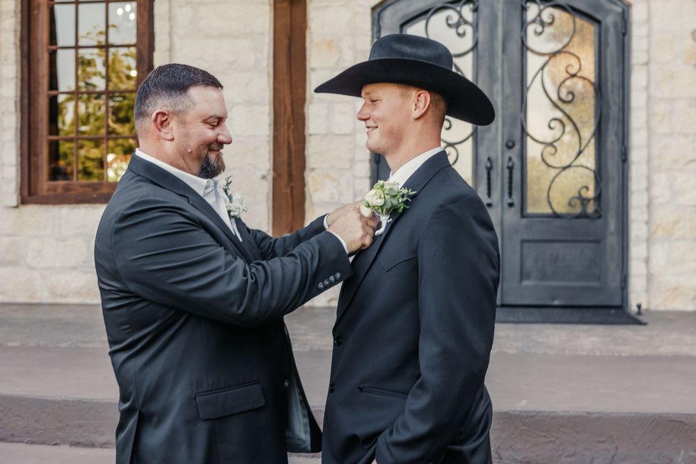 Two men in formal attire stand together outdoors while adjusting a tie and boutonniere before a special event.
