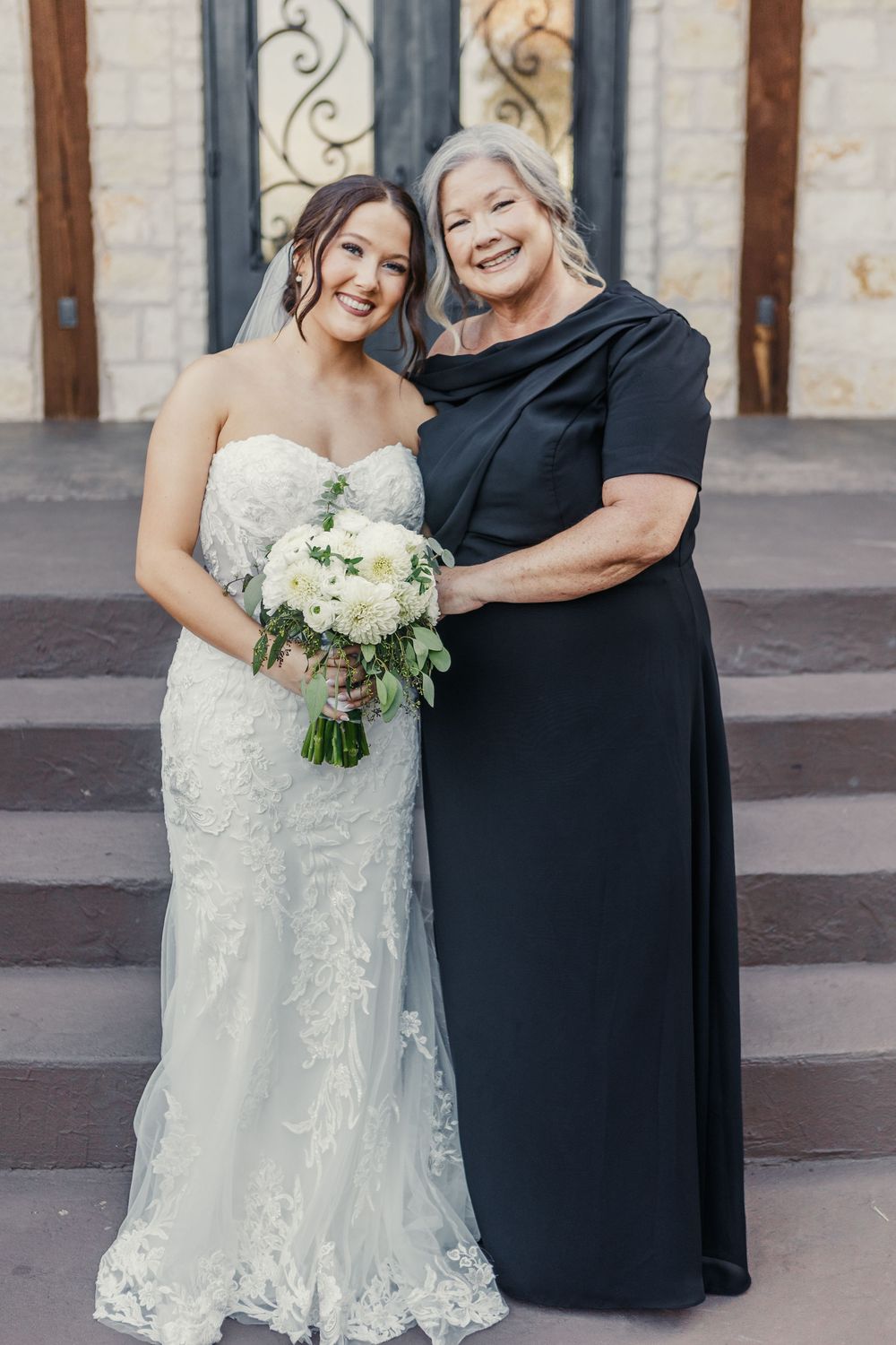 Bride in a white lace dress shares joyful moments with someone wearing black in an outdoor setting.