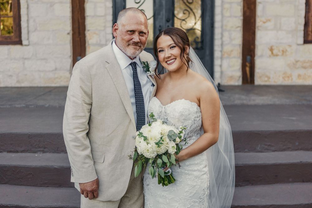 A bride in a white strapless dress holds a bouquet while posing with someone in a beige suit outside a church.
