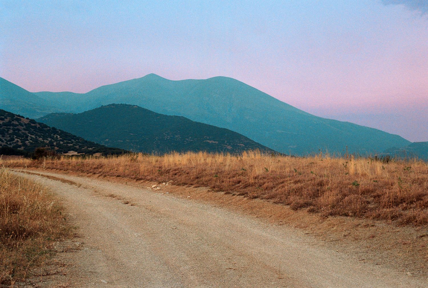 Dirt road leading toward mountains under soft evening sky in a rural landscape.