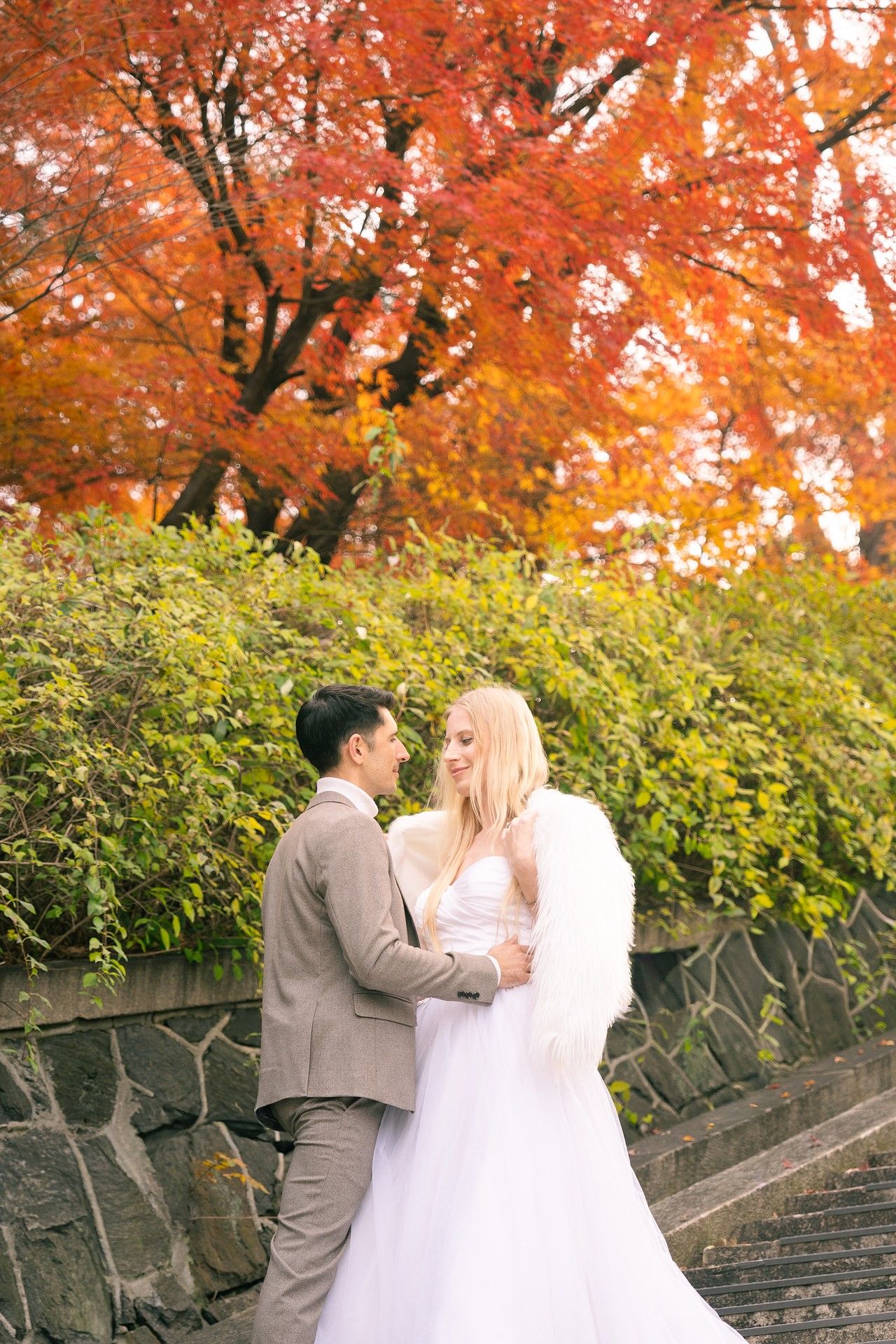 A couple in wedding attire shares an intimate moment under vibrant orange autumn foliage in a garden setting.