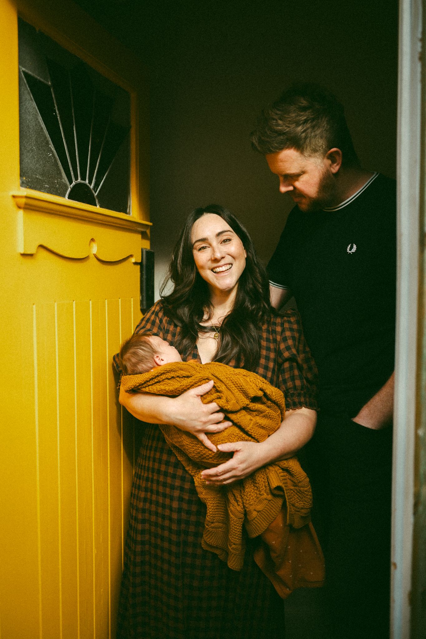 Two adults sitting in a cozy living room with artwork on sage green walls while holding an infant in mustard yellow clothing.