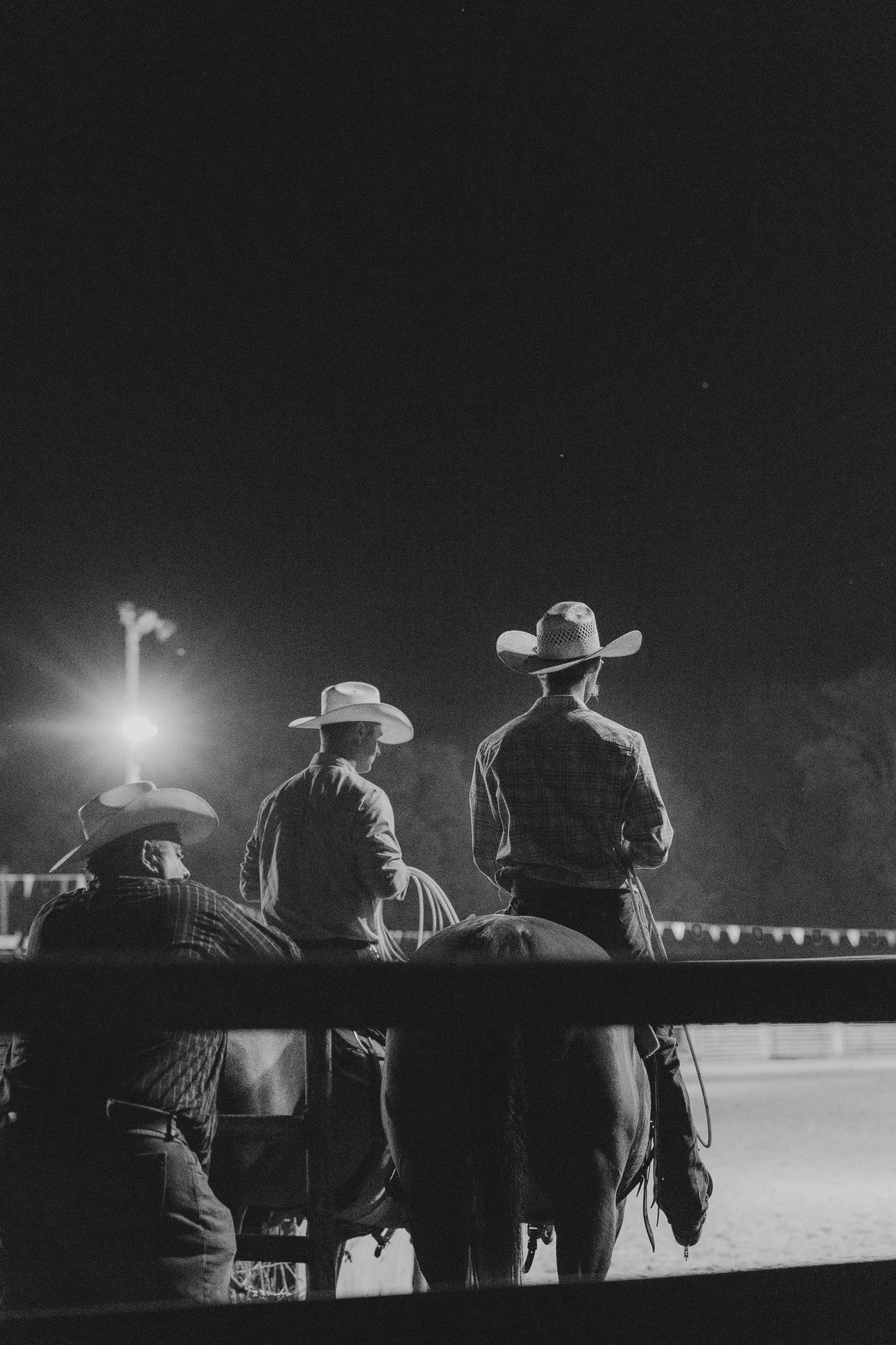 Two cowboys in cowboy hats lean against a fence at night while watching rodeo arena activities in black and white.