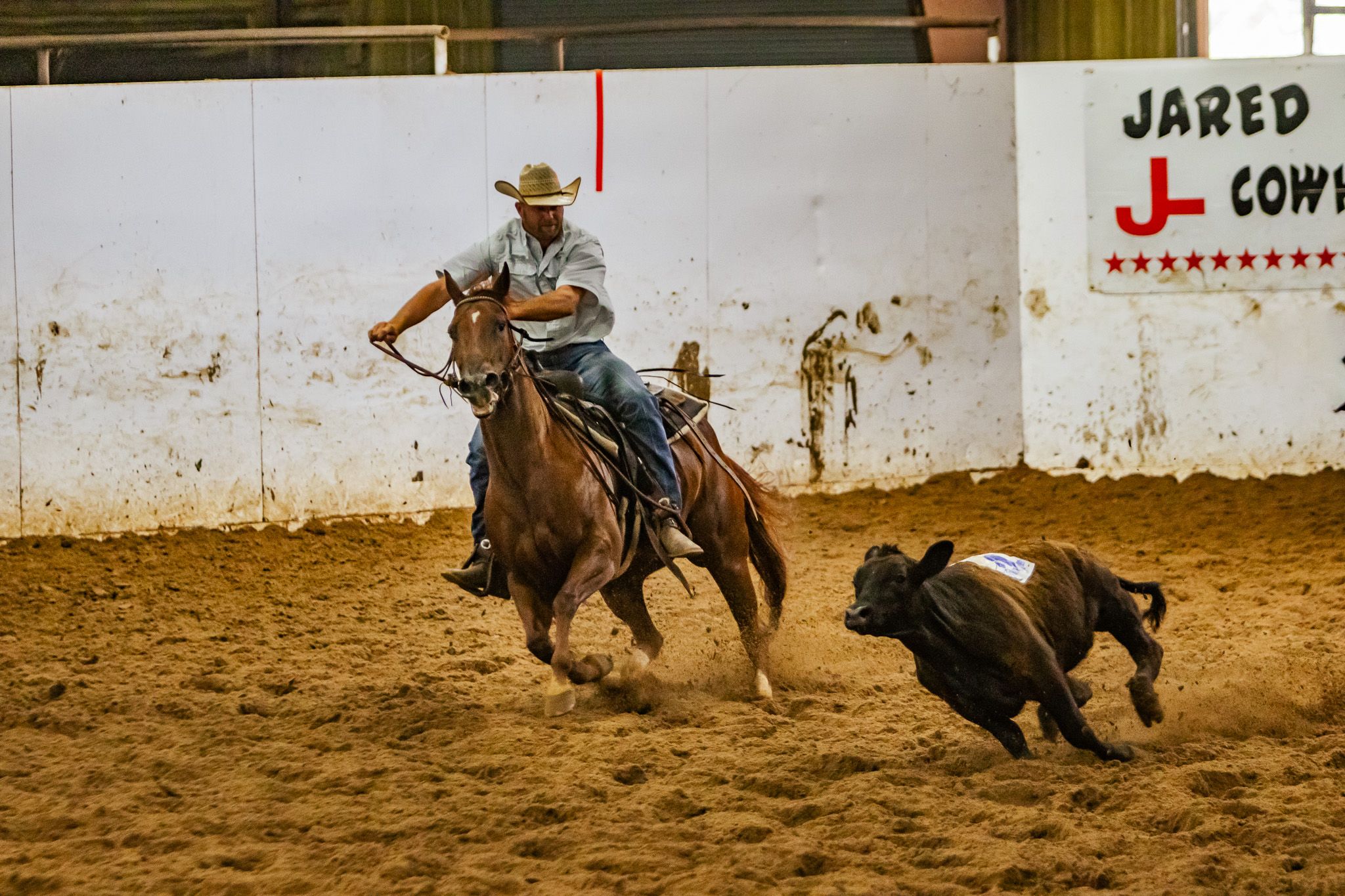 A cowboy on horseback competes in a rodeo calf roping event inside a dusty indoor arena.