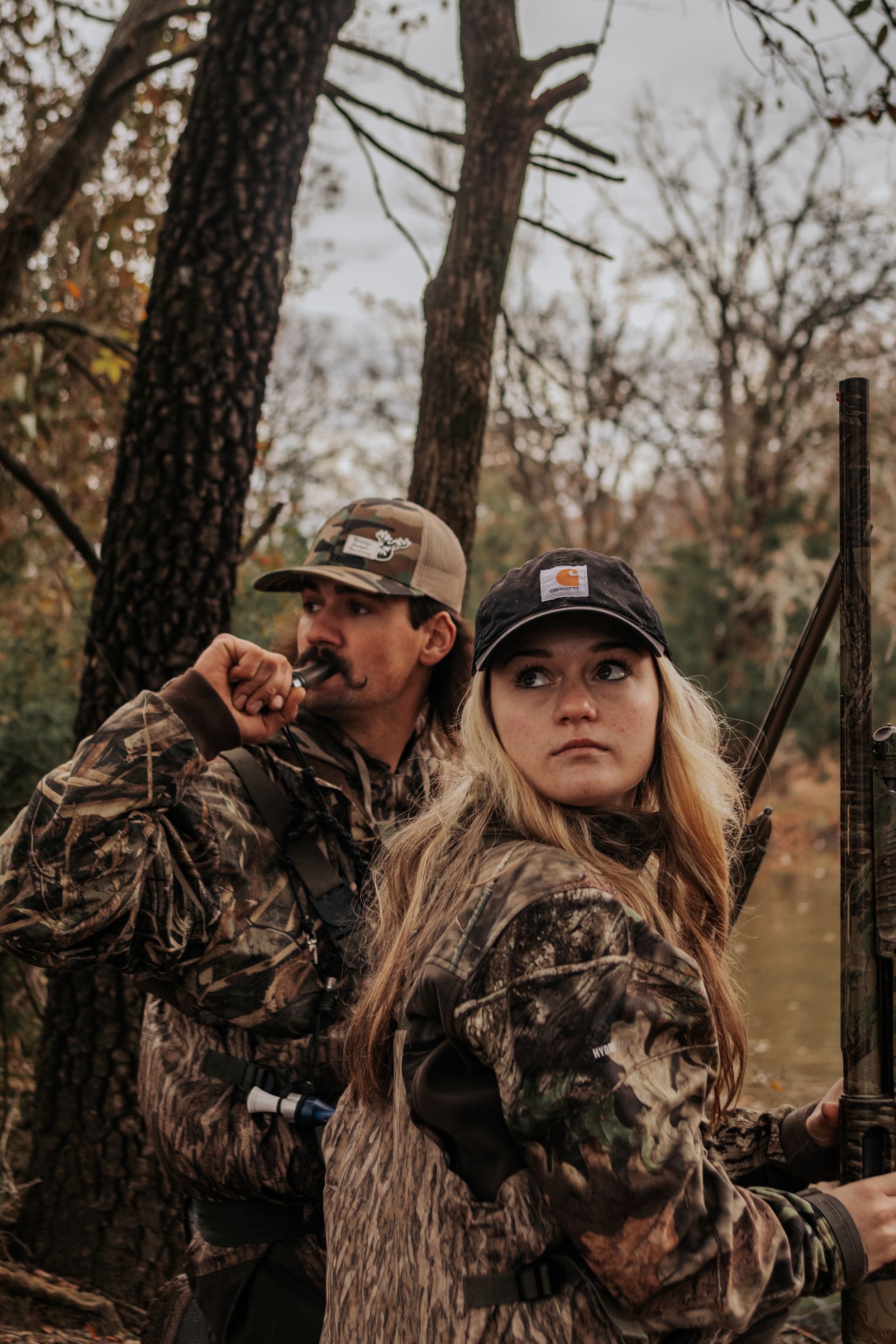 Two hunters in camouflage gear stand among trees during hunting season with autumn foliage in the background.