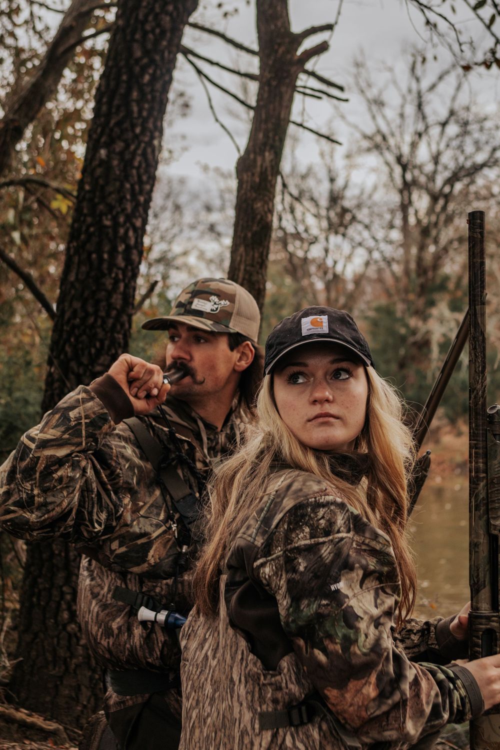 Two hunters in camouflage gear stand among trees during hunting season with autumn foliage in the background.