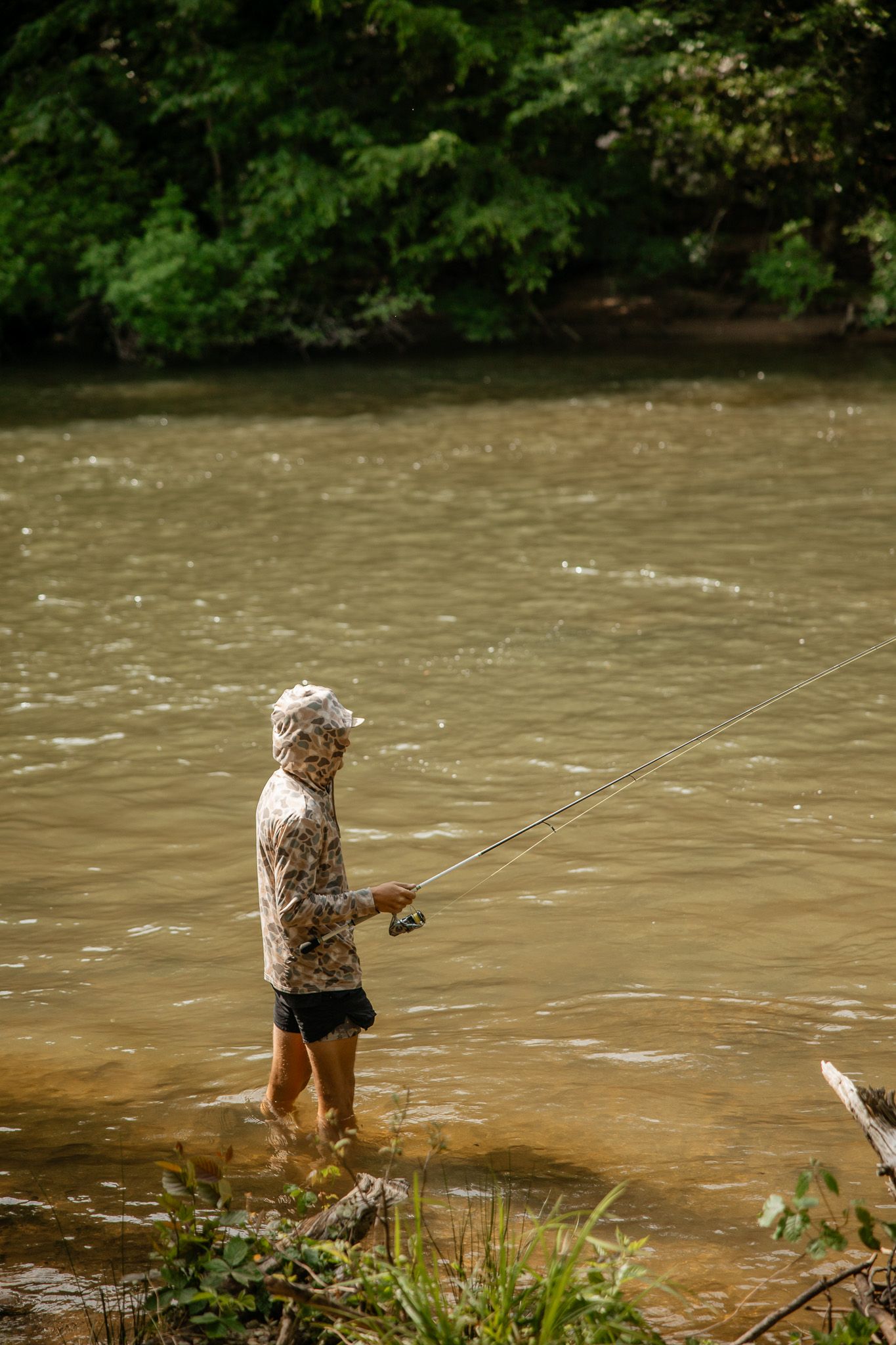 An angler wearing floral attire wades in muddy brown river water while fishing near a wooded shoreline.