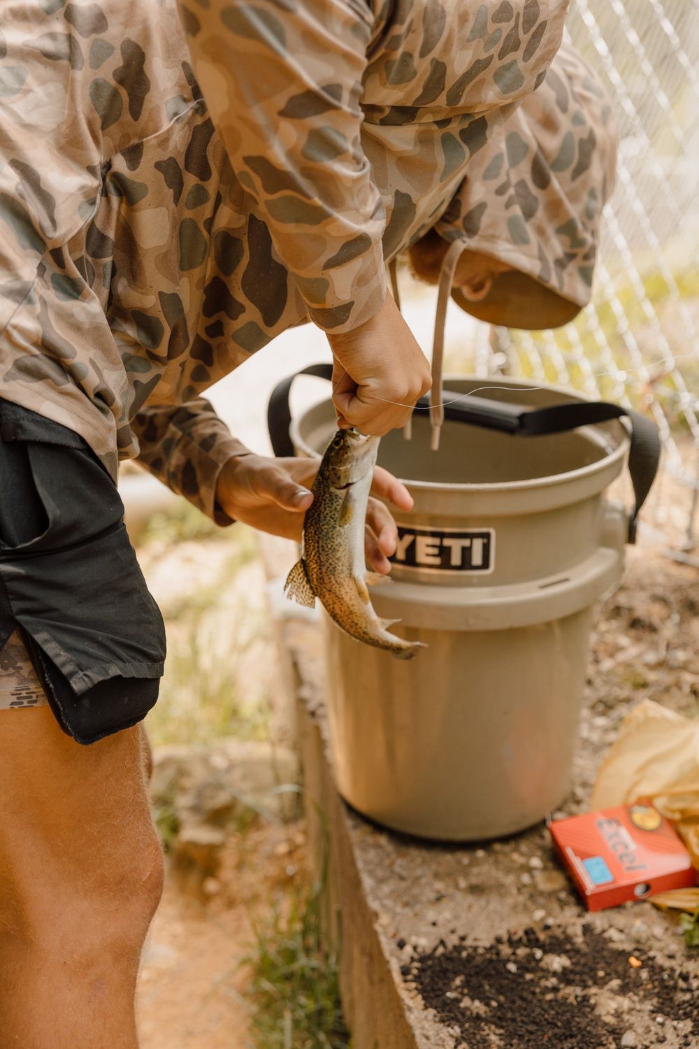 A person holds a freshly caught trout over a tan YETI bucket during an outdoor fishing activity.