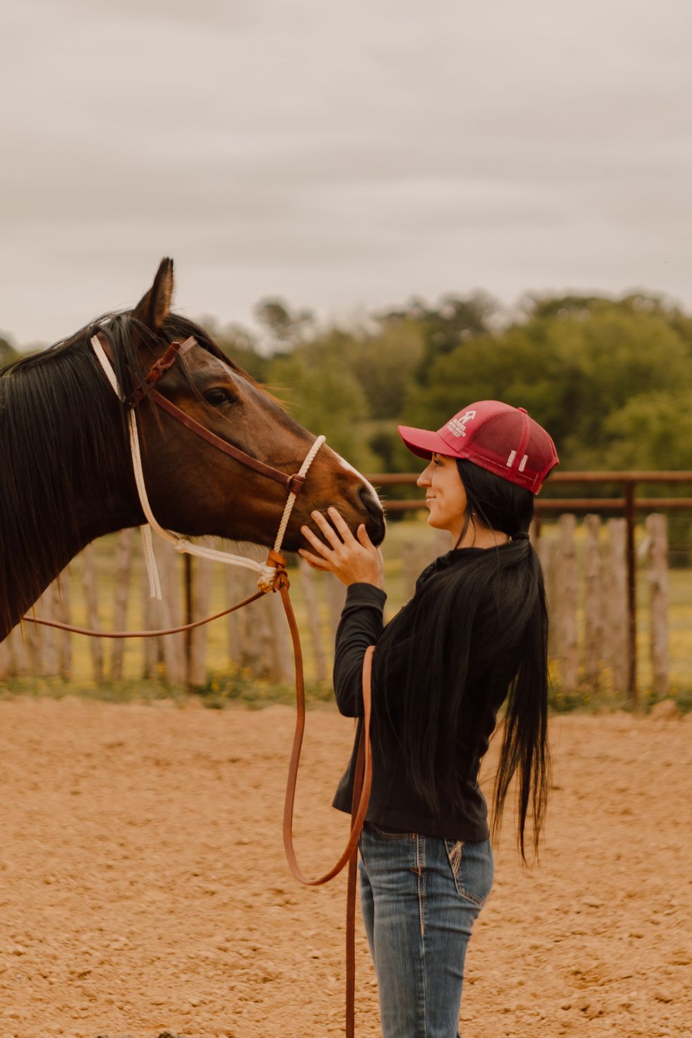 A horse and rider share a gentle moment in a dusty arena during sunset as the rider pets the horse's nose.
