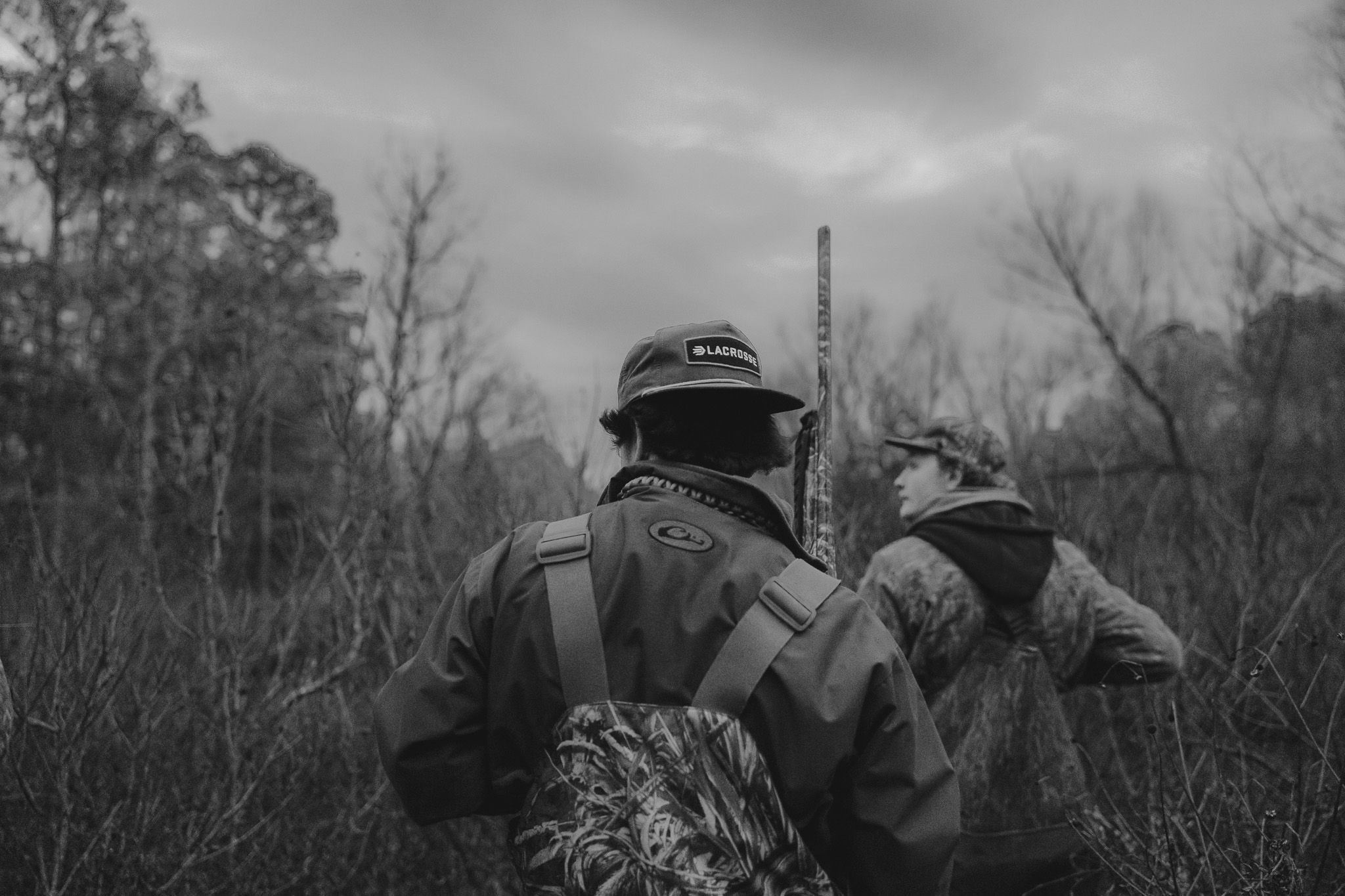 Two hunters in camouflage gear crouching in tall grass during an overcast day in black and white.