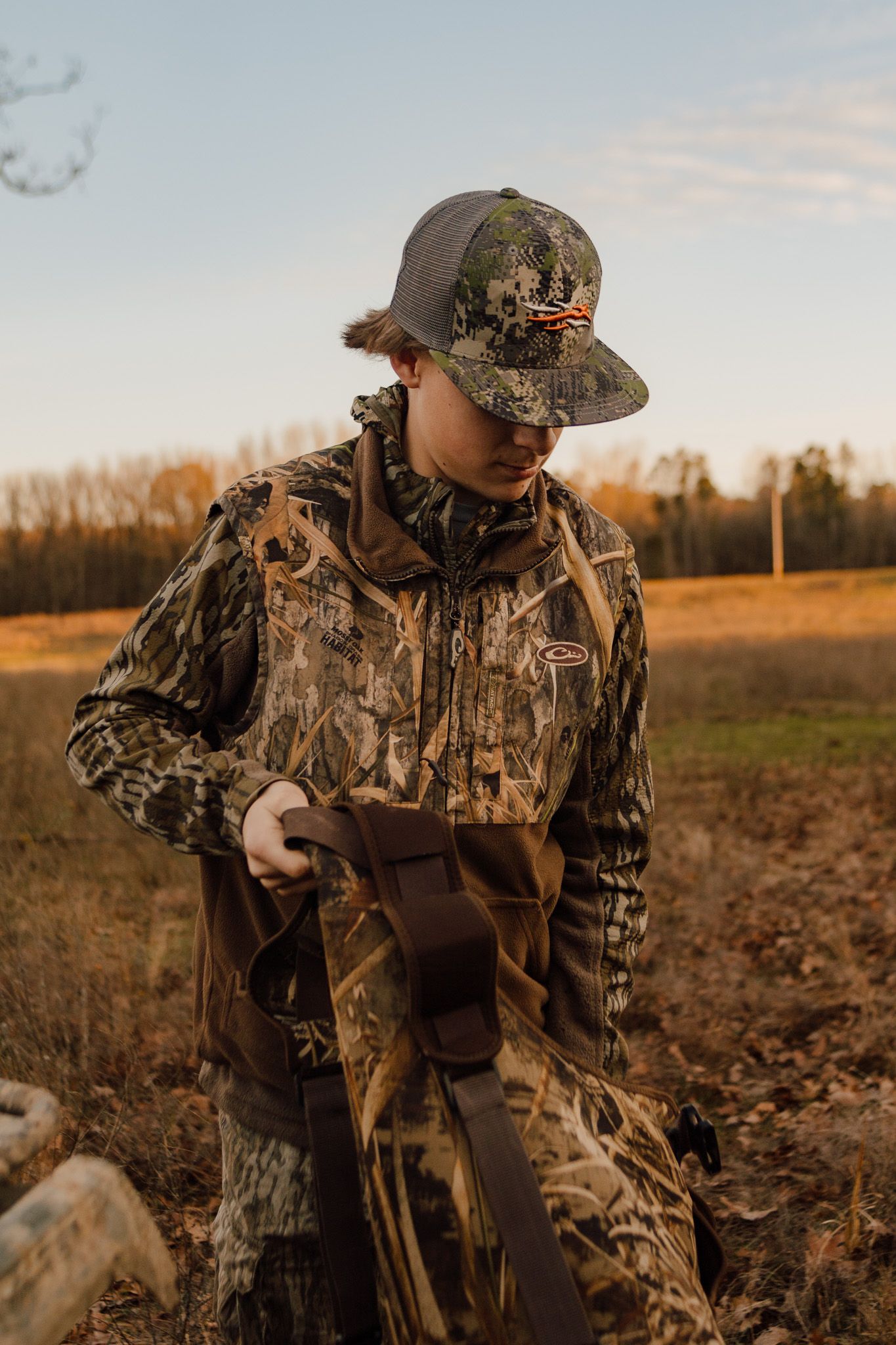 Hunter wearing camouflage gear and cap stands in grassy field during golden hour sunset.
