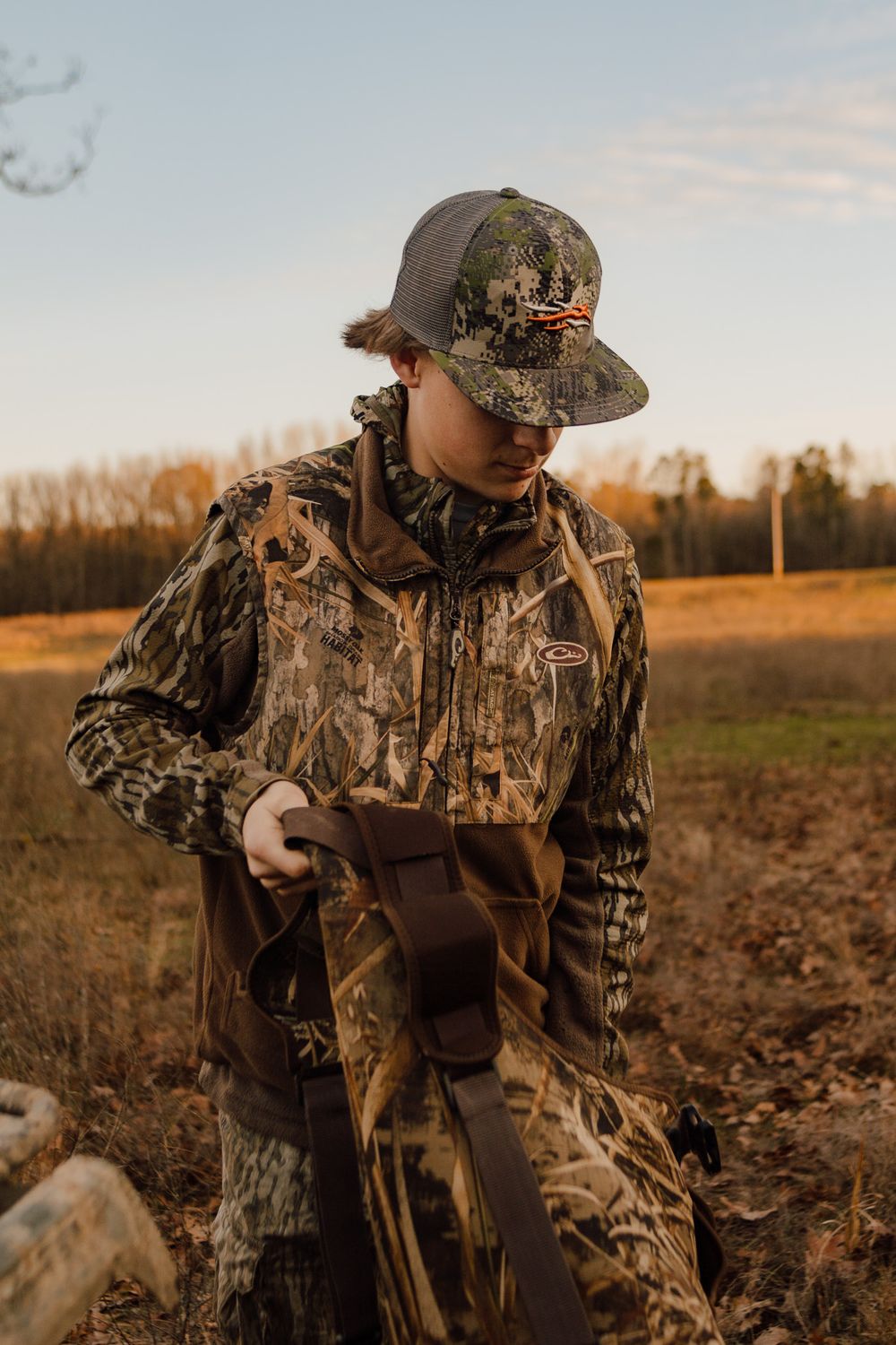 Hunter wearing camouflage gear and cap stands in grassy field during golden hour sunset.