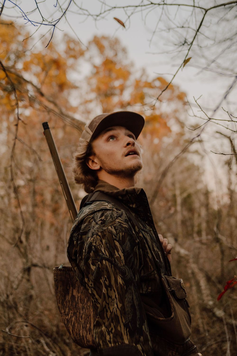 A hunter in camouflage gear scans the sky attentively in an autumn forest scene with dried leaves and bare branches.