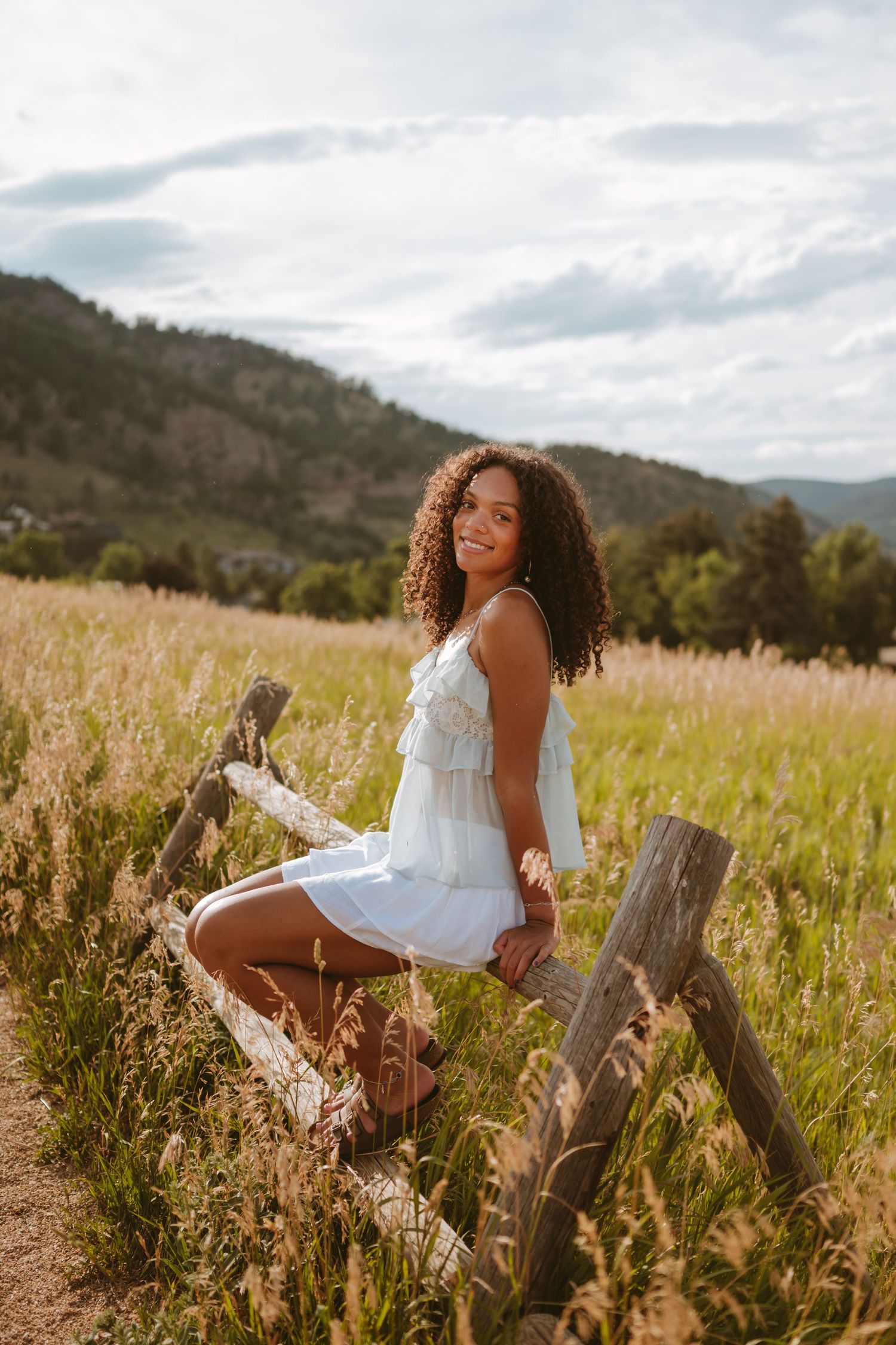 Person in flowing white dress sits on rustic wooden fence surrounded by golden meadow and mountain landscape at sunset.