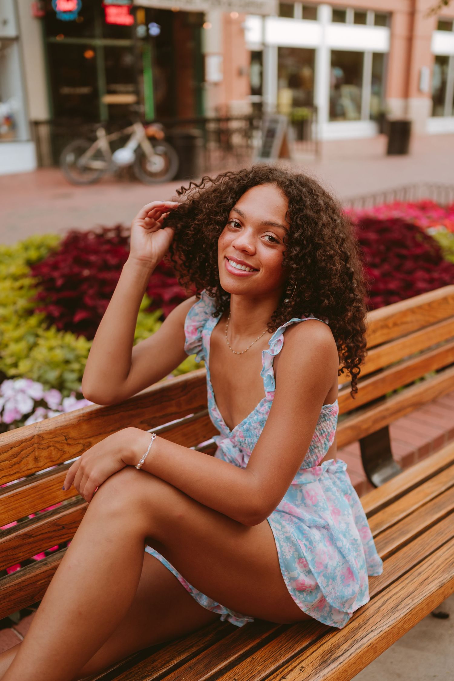 Woman with curly hair in floral sundress sits smiling on wooden bench near colorful flower garden.