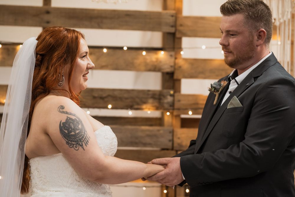 A bride and groom hold hands during their rustic barn wedding ceremony with wooden pallet backdrop.