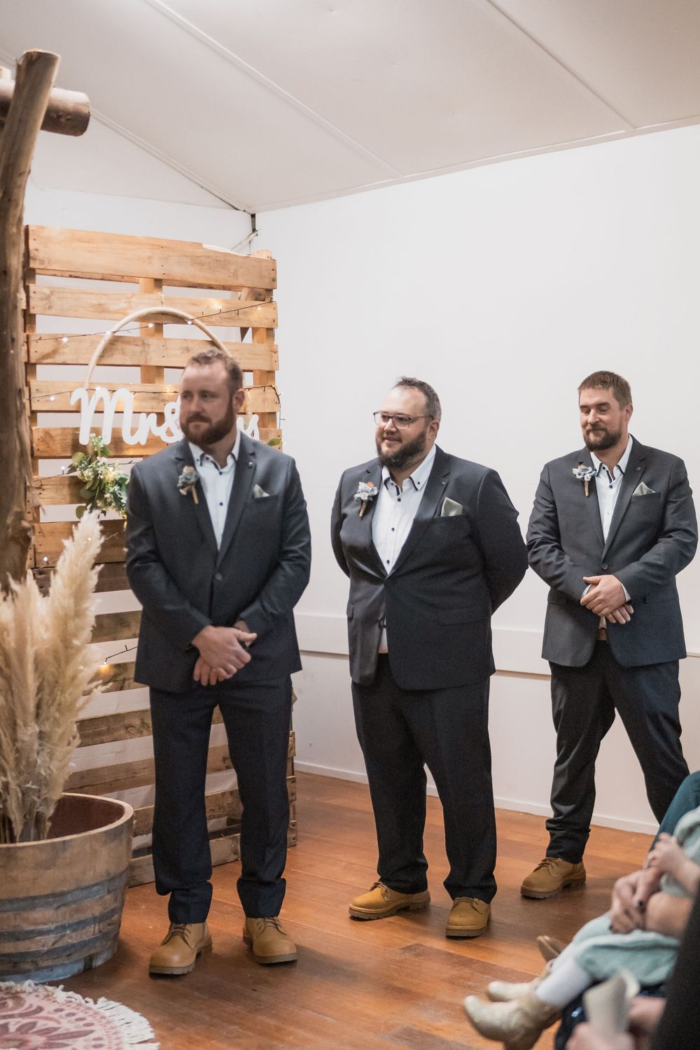 A series of four wedding party photos shows groomsmen in black suits posing on dark hardwood floors at a venue.