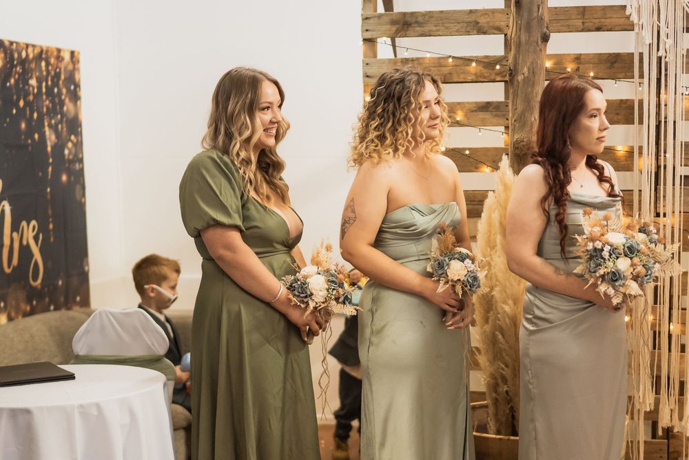 Group of women in different shades of olive green dresses standing next to wooden backdrop during formal event.