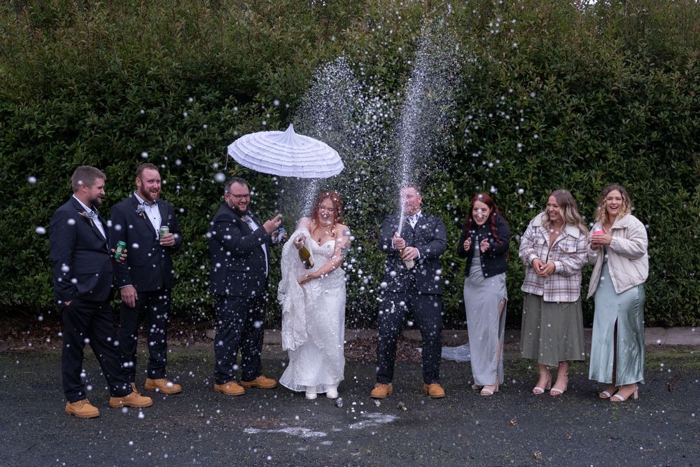 Wedding party standing together under an umbrella as soap bubbles or confetti falls around them outdoors.