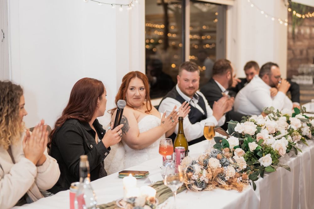 Wedding guests applaud during reception dinner at elegant venue with floral centerpieces and string lights.