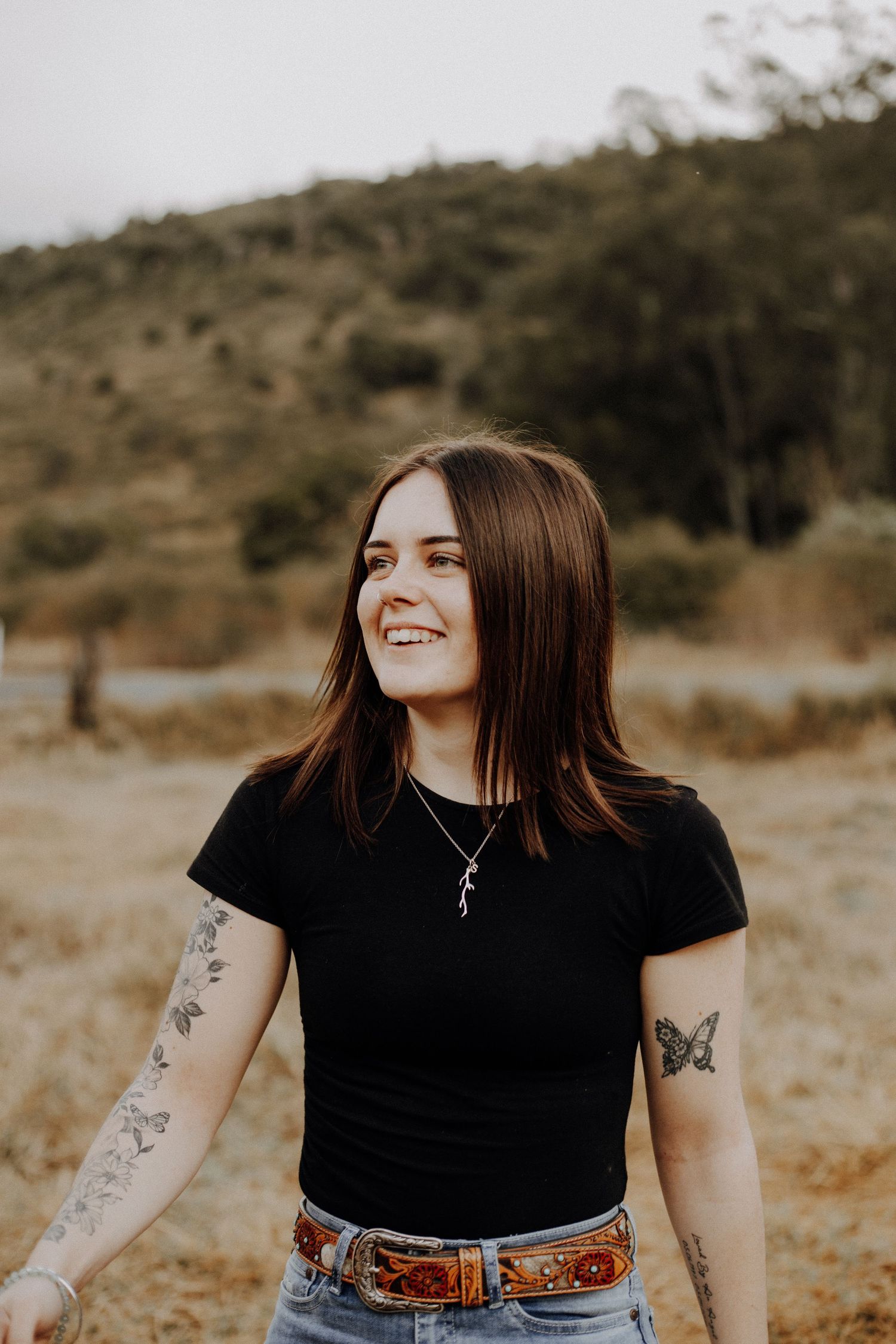 A young person in a black t-shirt and jeans smiles outdoors against a natural landscape with visible arm tattoos.