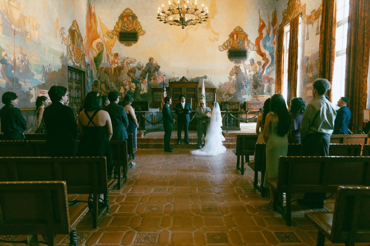 A traditional church wedding ceremony with guests seated in wooden pews under ornate painted walls and a chandelier.