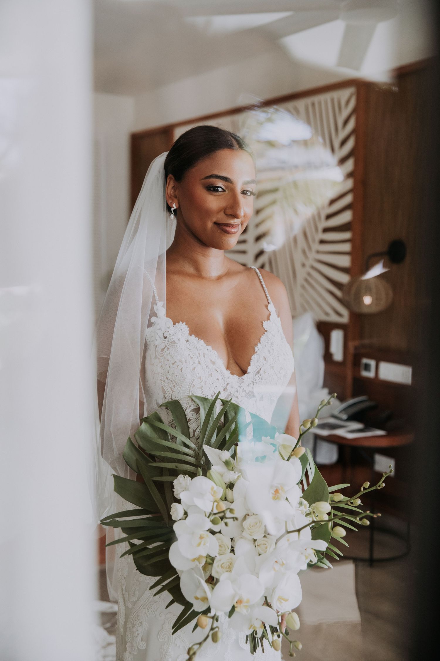 Bride holding white orchid bouquet poses in lace wedding dress with tropical palm leaf accent.