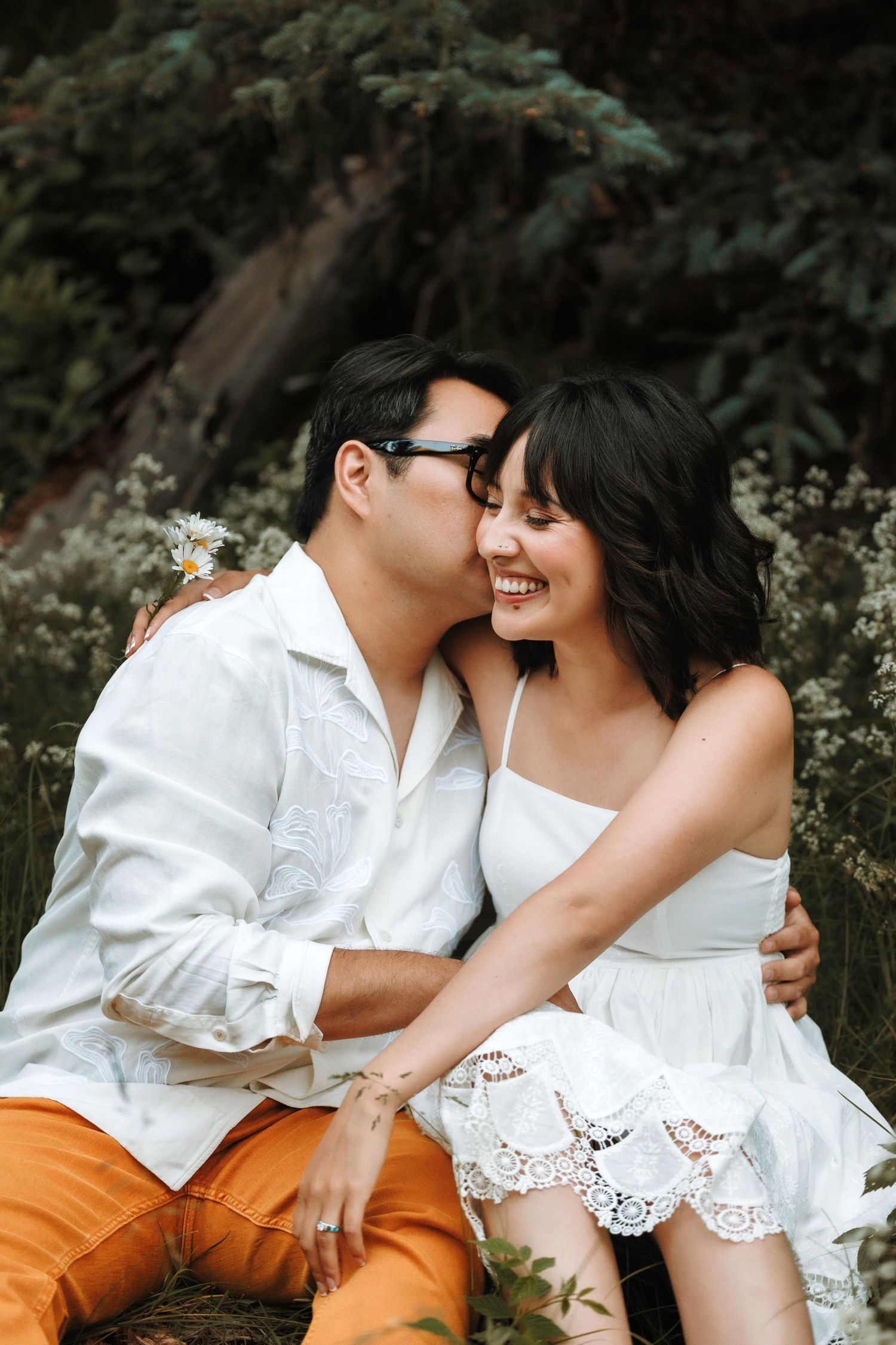 Smiling couple sits together among wildflowers as man whispers in womans ear outdoors.