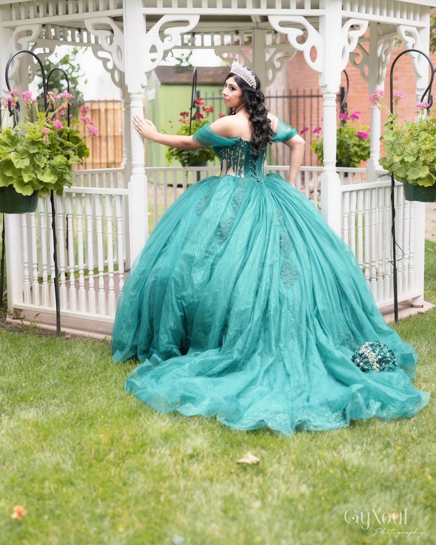 A stunning teal ballgown with off-shoulder details is showcased in front of a white garden gazebo with hanging plants.