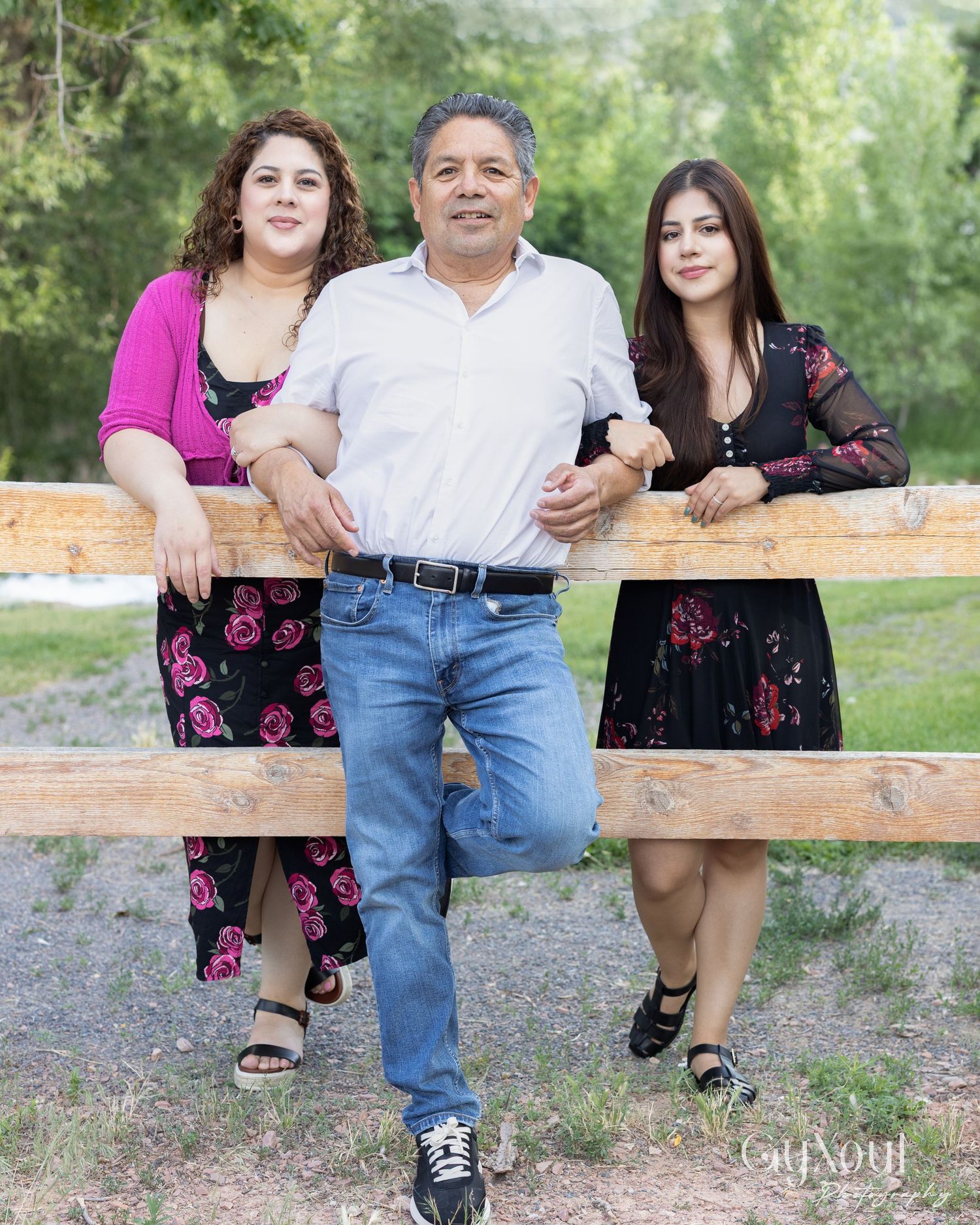 A family group in coordinating floral dresses and casual attire poses together by a wooden fence in an outdoor setting.