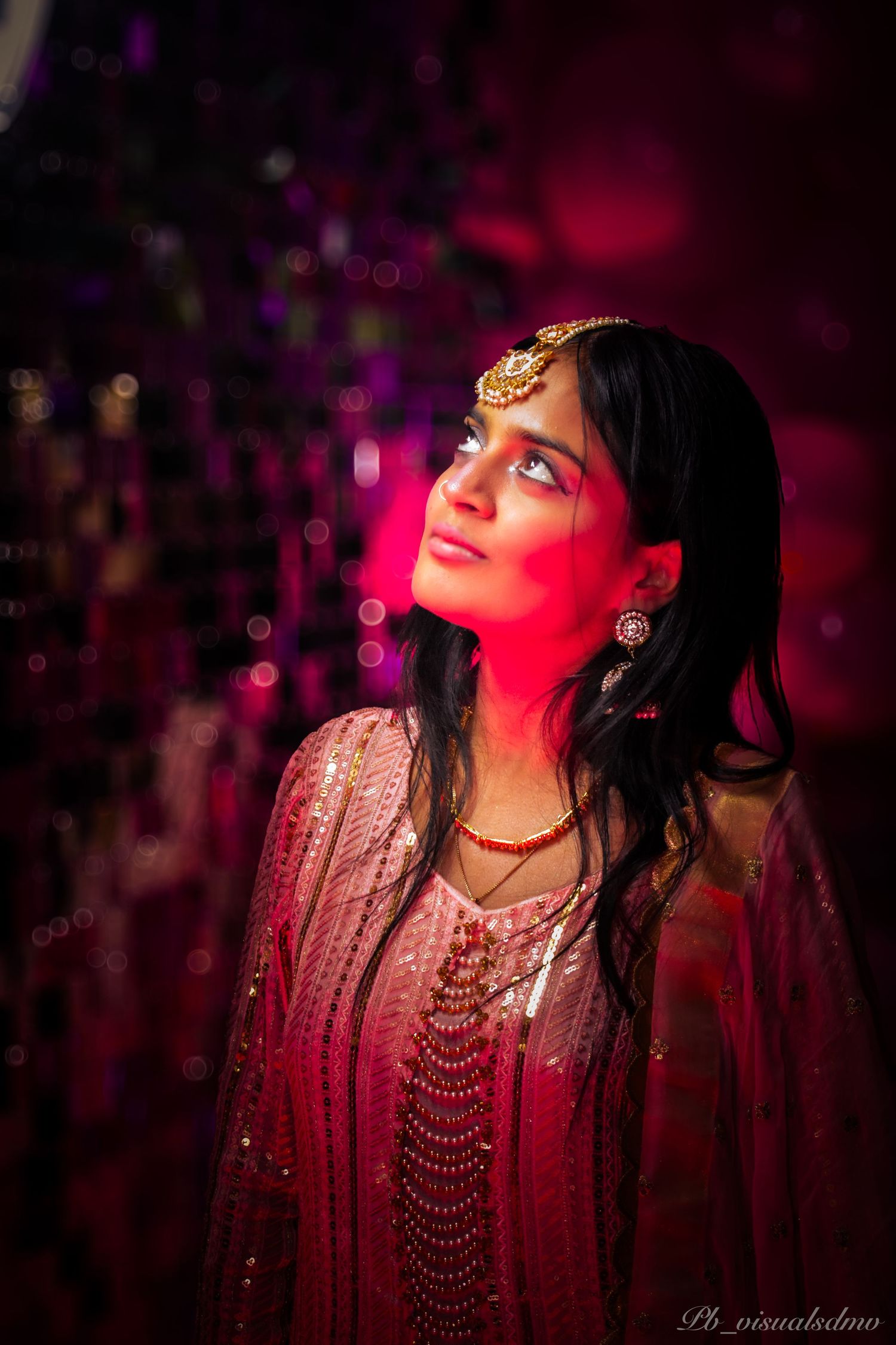 Our model in traditional South Asian attire with gold jewelry, looking upward under dramatic pink lighting during a celebration.