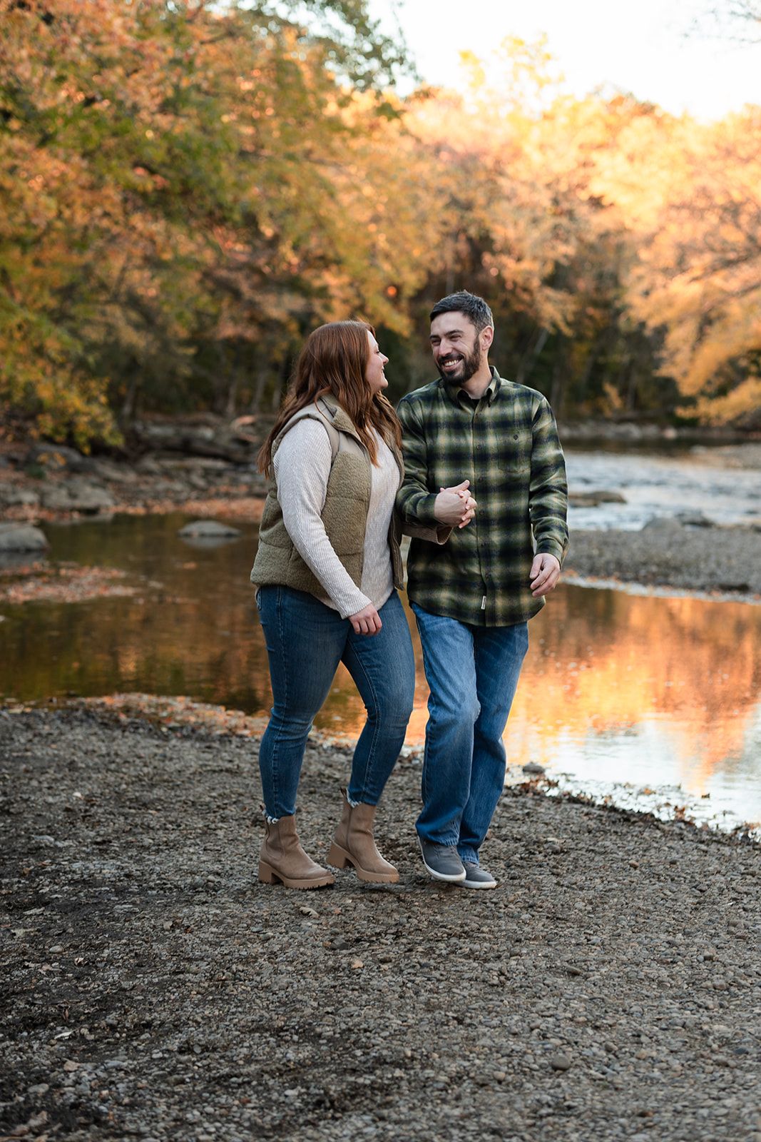 A couple laughs while enjoying a romantic autumn walk along a creek with vibrant fall foliage reflected in the water.