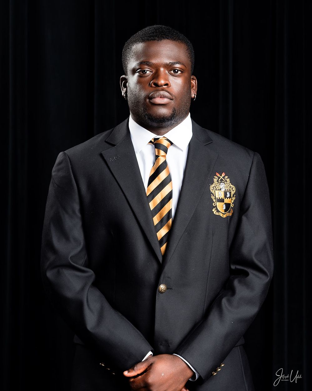 Formal portrait of student in dark suit with striped tie and school emblem against black background.
