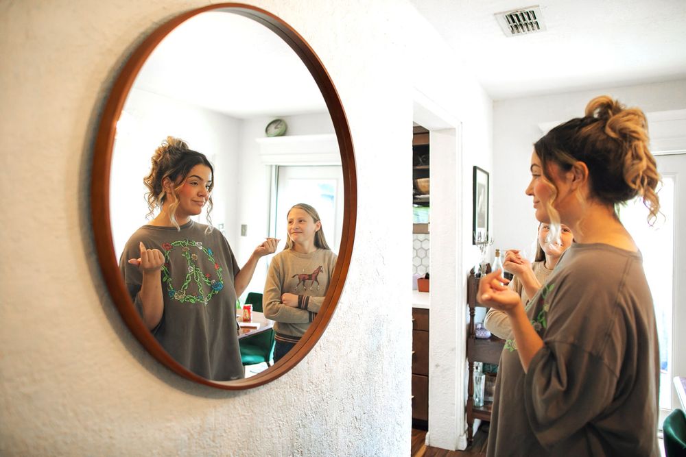 Two women prepare in front of a large round mirror hanging on a wall while getting ready for a special event.