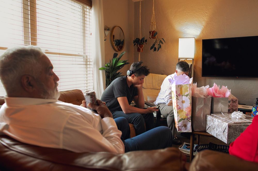 Healthcare workers attend to a patient during a home health visit in a living room with natural light coming through window blinds.